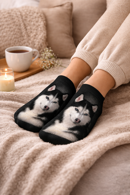 A person wearing black crew socks with a printed husky face sits on a beige blanket with a cup of coffee, a candle, and pillows in the background.