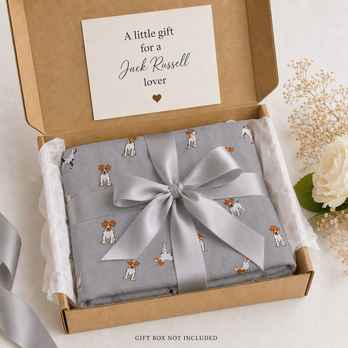 A light grey scarf gift wrap with a pattern of small Jack Russell Terrier dogs, wrapped in a wide silver satin ribbon and tied in a large bow, presented inside an open brown kraft gift box with a white card that reads 'A little gift for a Jack Russell lover'.
