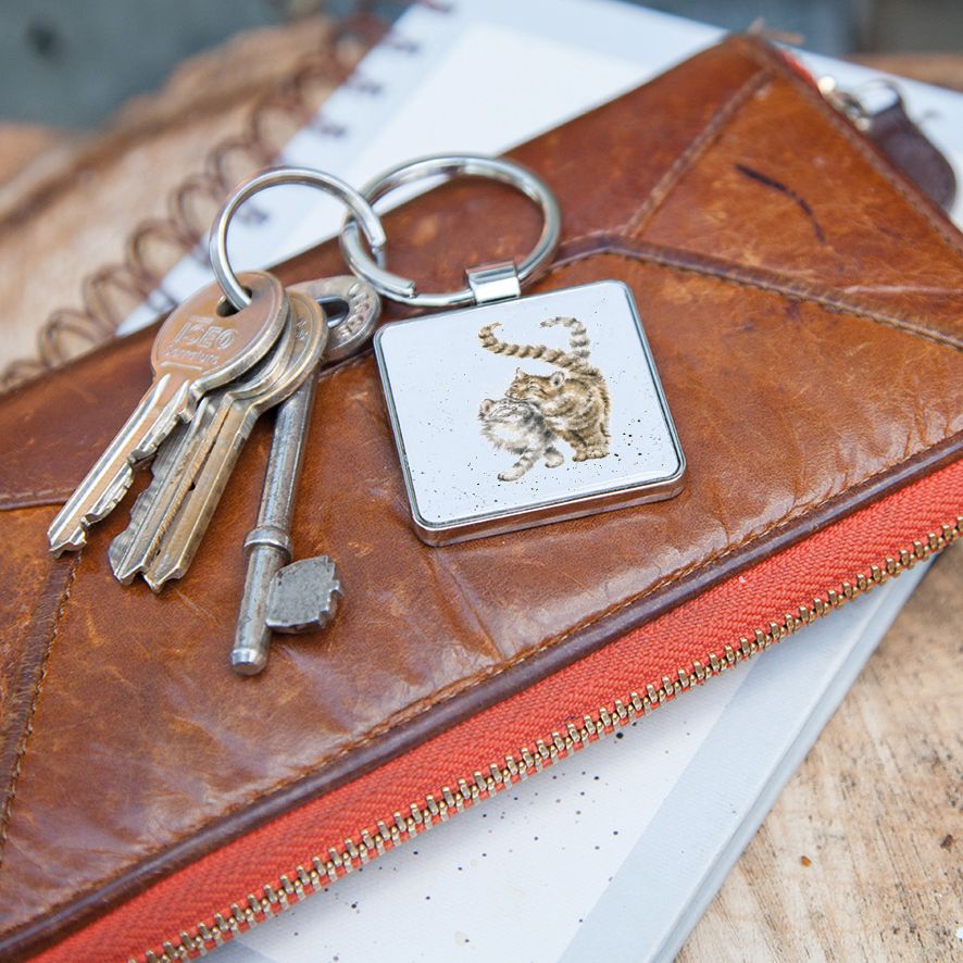 A square silver keychain with a raised cat design, shown on a brown leather zip pouch with several keys, resting on a white notebook over a wooden surface.