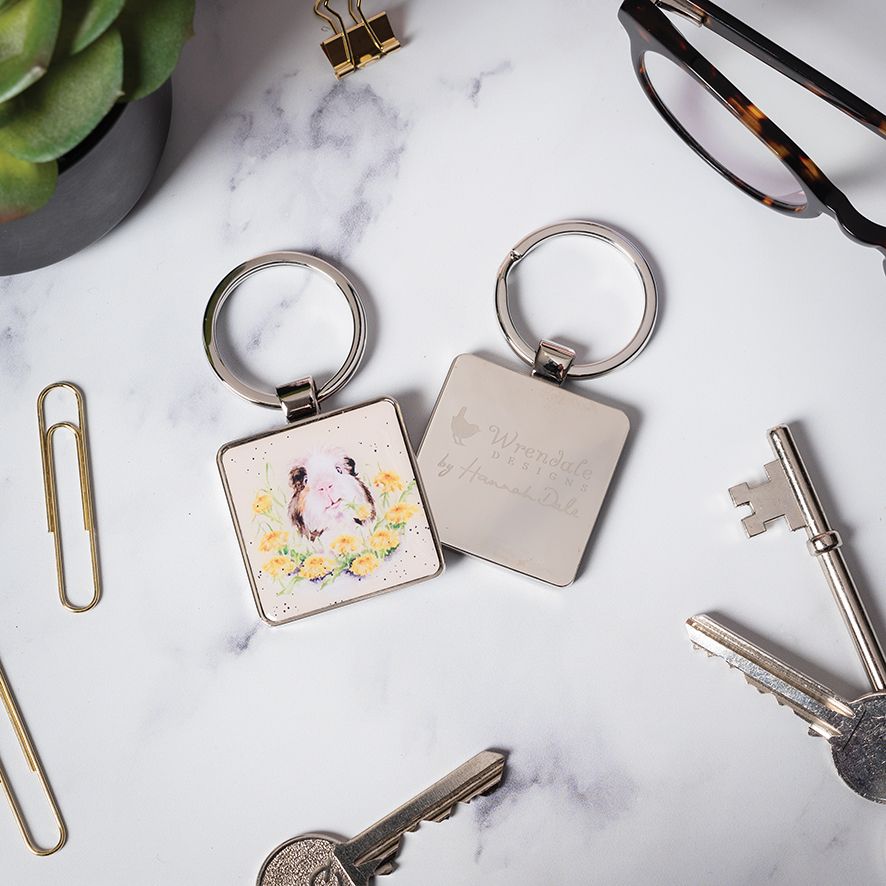 Two square metal keychains on a white marble surface with office props. One keychain has a silver ring and a white square charm with a watercolor-style illustration of a cat and yellow flowers.
