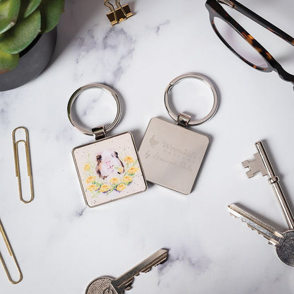 Two square metal keychains on a white marble surface with office props. One keychain has a silver ring and a white square charm with a watercolor-style illustration of a cat and yellow flowers.