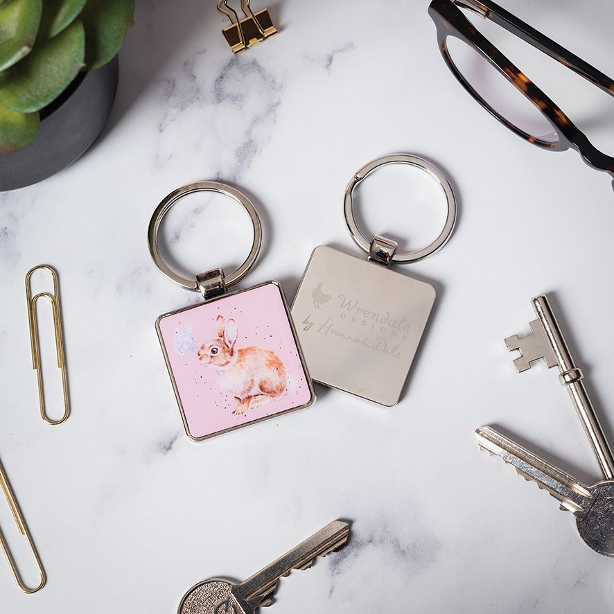 Two square metal keychains on a white marble surface with office props. One keychain has a pink background with a watercolor rabbit illustration, and the other is silver with engraved text.