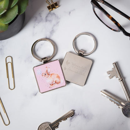 Two square metal keychains on a white marble surface with office props. One keychain has a pink background with a watercolor rabbit illustration, and the other is silver with engraved text.