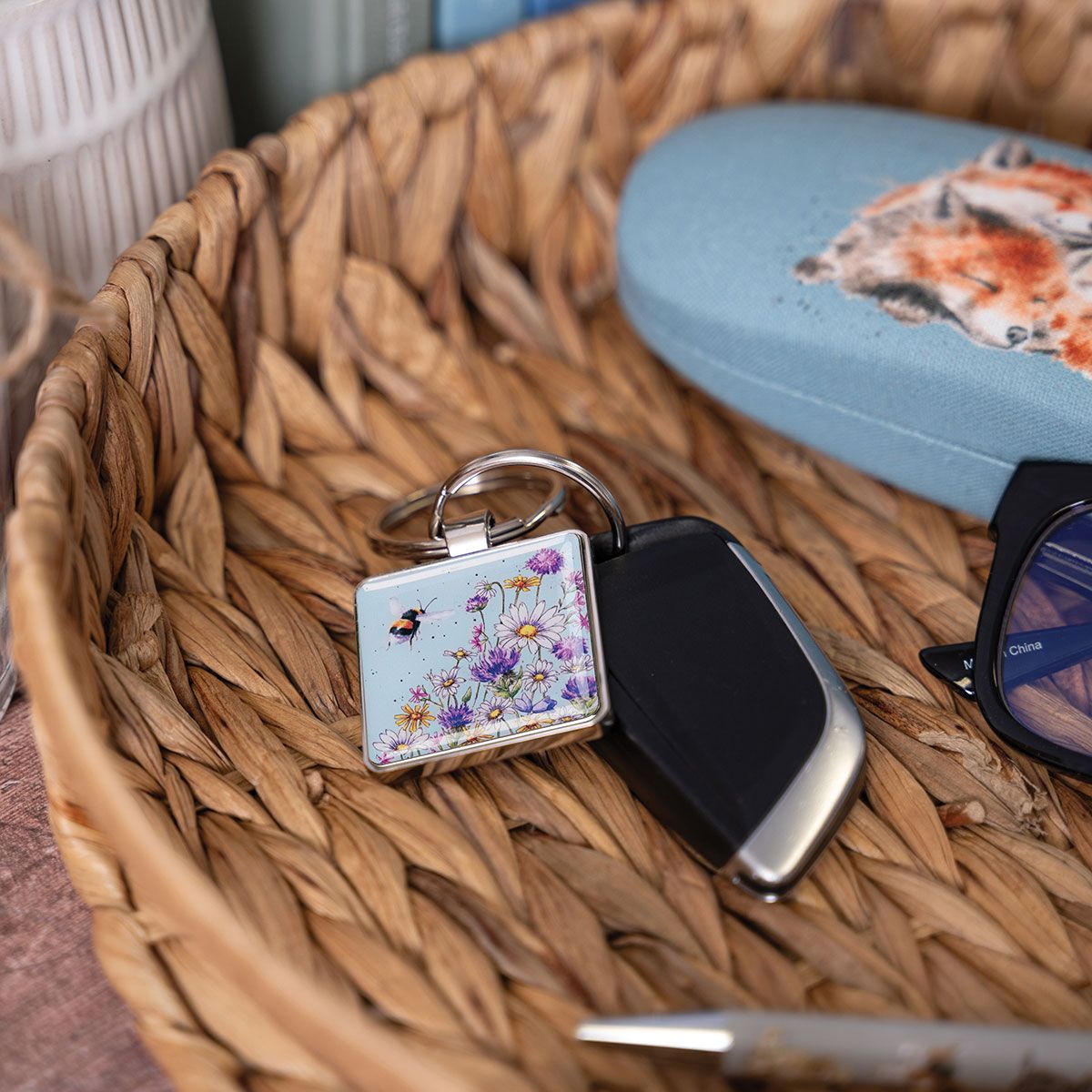 A square keychain with a floral and bee design attached to a black car key fob, resting inside a woven wicker basket next to sunglasses and a blue case.