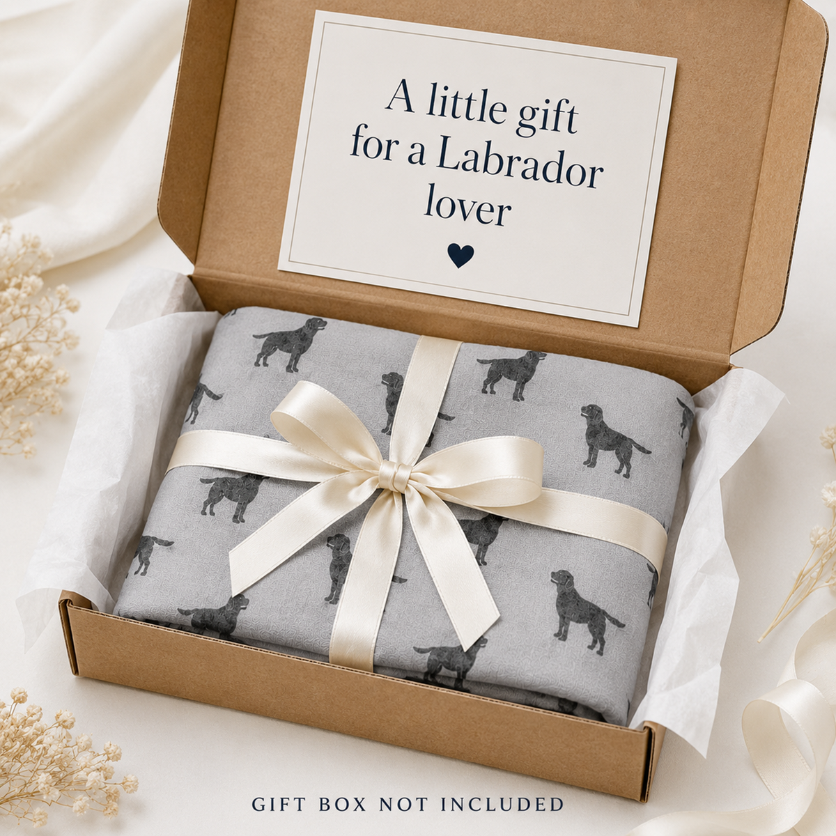 A folded grey scarf gift wrapped with a black Labrador dog print pattern, tied with a cream satin ribbon and presented in a brown kraft gift box. The inside lid has a printed card that reads 'A little gift for a Labrador lover'.