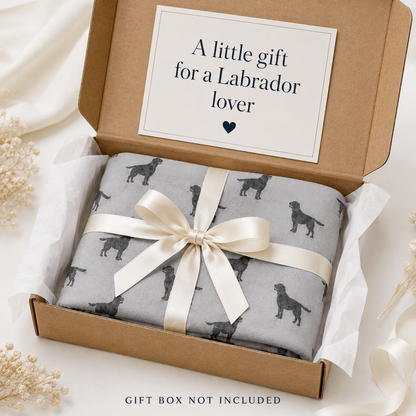 A folded grey scarf gift wrapped with a black Labrador dog print pattern, tied with a cream satin ribbon and presented in a brown kraft gift box. The inside lid has a printed card that reads 'A little gift for a Labrador lover'.
