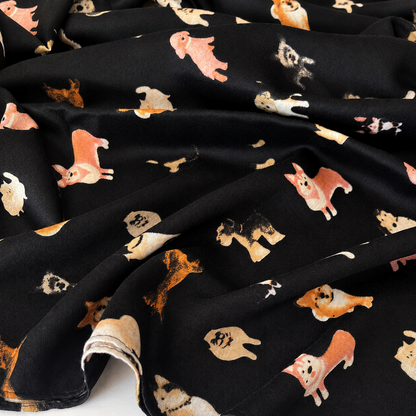 A folded black scarf featuring a playful print of various dog breeds in black, white, beige, and orange, finished with fringed edges.