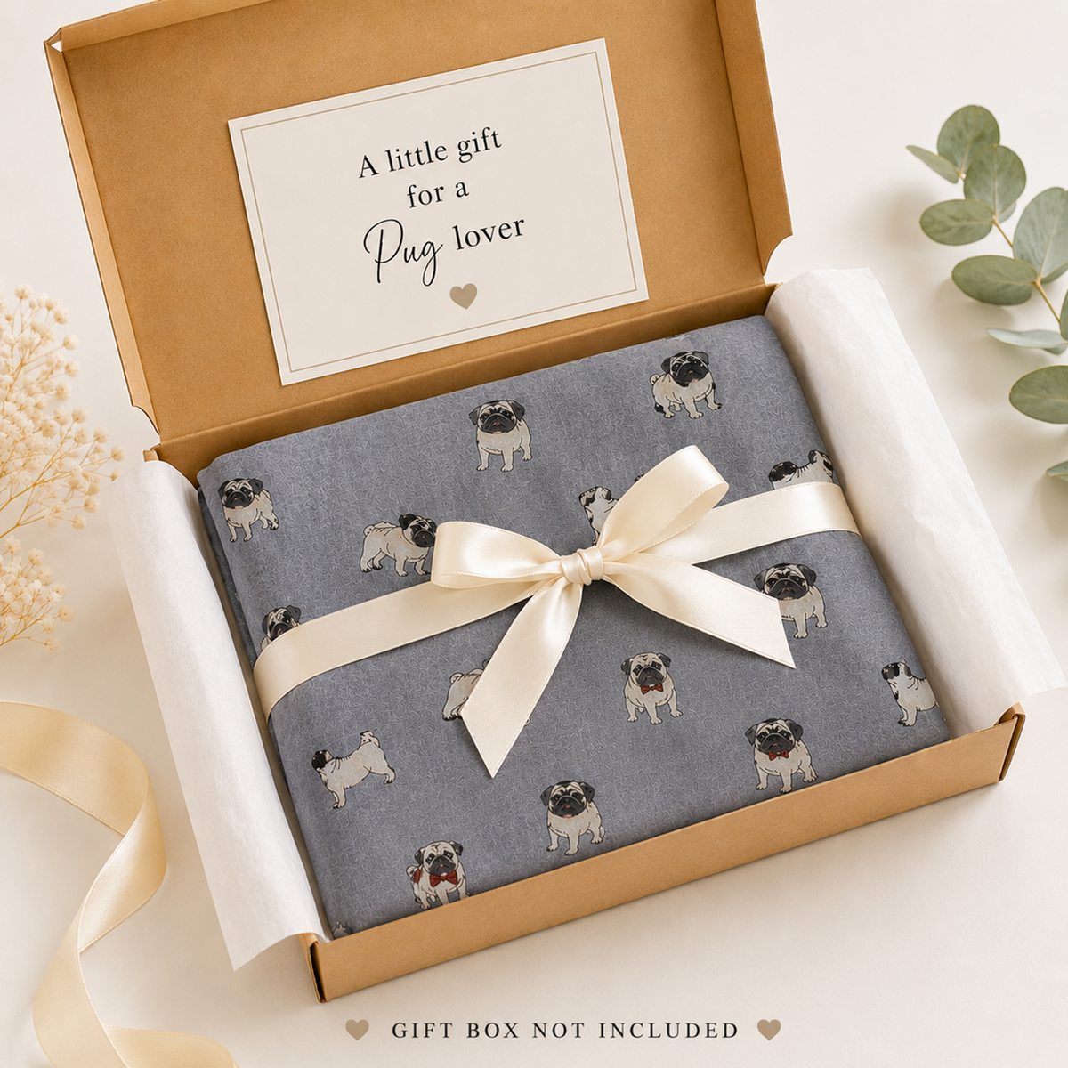 A gift-wrapped product box shown from above on a light neutral background. The scarf is grey with a repeating pattern of small pug dogs, tied with a cream satin ribbon and bow.