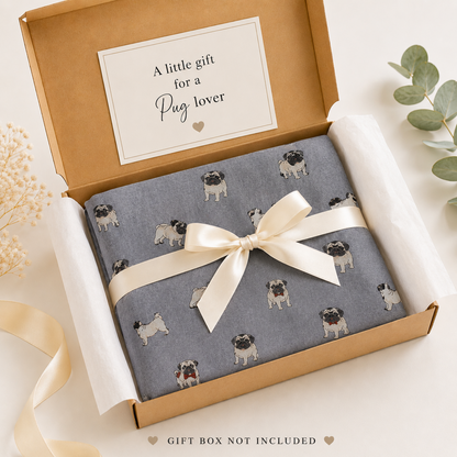 A gift-wrapped product box shown from above on a light neutral background. The scarf is grey with a repeating pattern of small pug dogs, tied with a cream satin ribbon and bow.