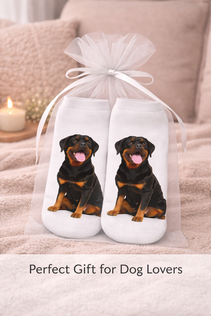 A pair of white socks with printed Rottweiler dog images, tied in a clear tulle gift bag with a white ribbon, set against a cozy bedroom background with a lit candle and the text 'Perfect Gift for Dog Lovers' at the bottom.