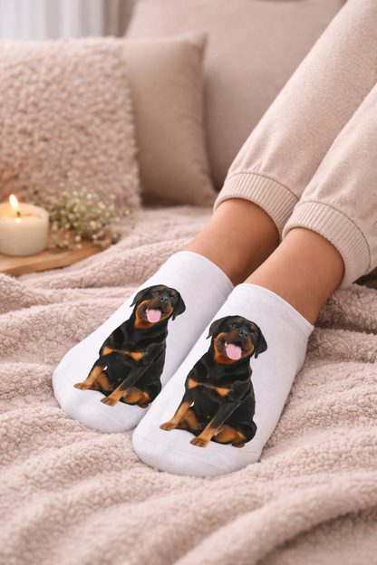 White low-cut socks featuring a printed Rottweiler dog design, worn on feet resting on a cozy beige blanket with a candle and pillows in the background.