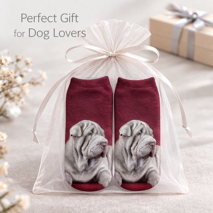 A pair of Shar Pei dog-themed trainer socks inside a clear tulle gift bag with a white ribbon, displayed on a neutral fabric surface with decorative dried flowers and a gift box in the background. Text reads 'Perfect Gift for Dog Lovers'.