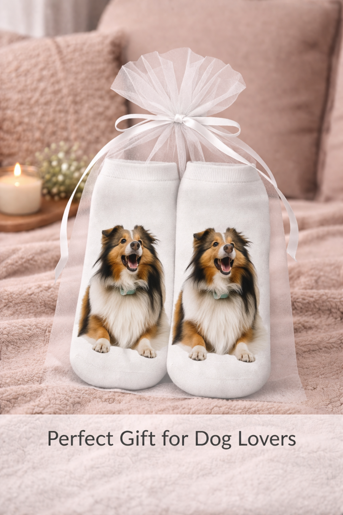 A pair of white socks with printed images of a happy Sheltie dog, tied together with a sheer white tulle bag and ribbon, set against a cosy bedroom background with a blanket and a lit candle. Text at the bottom reads 'Perfect Gift for Dog Lovers'.