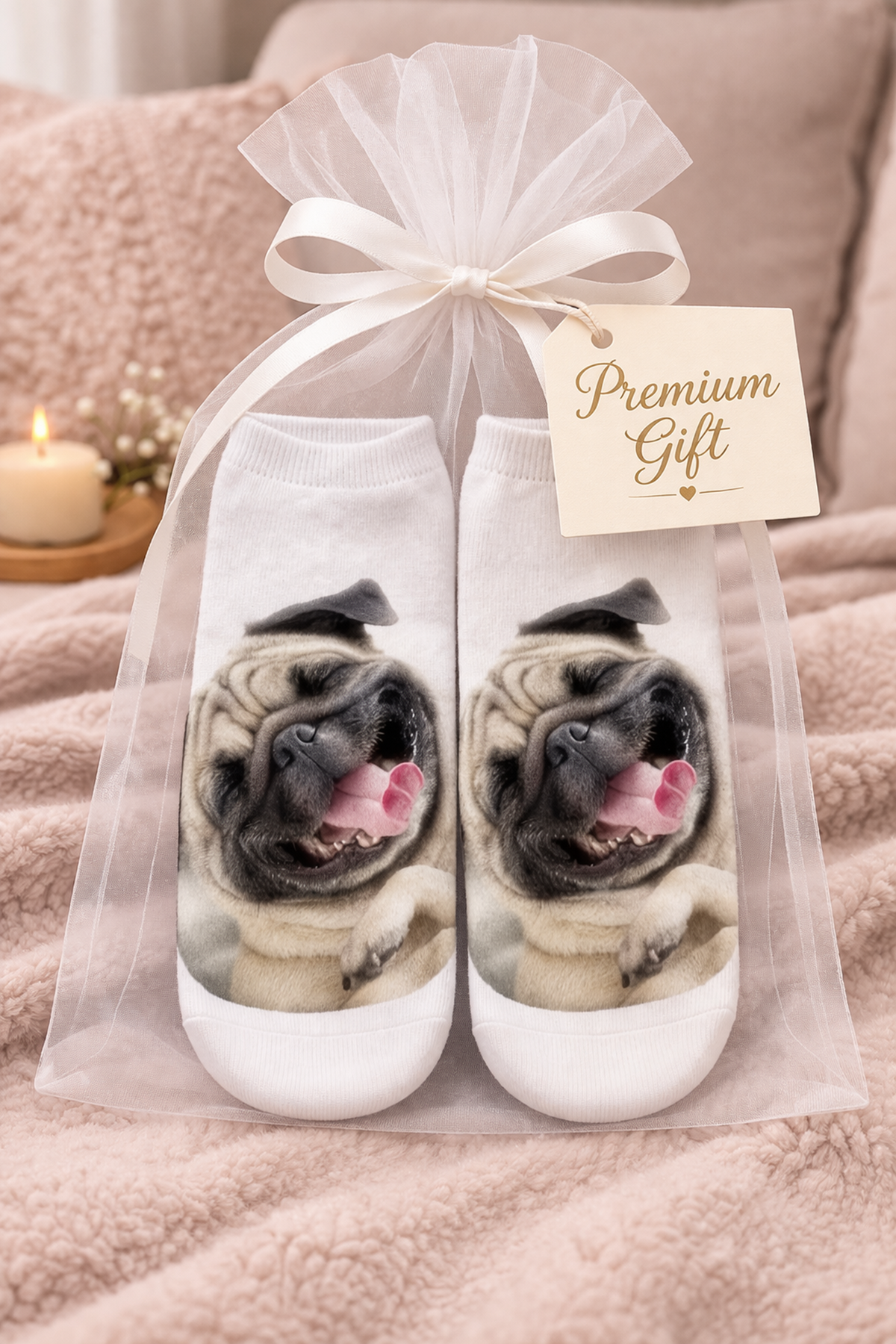 A pair of white socks with a printed pug dog face on each sock, presented in a clear tulle gift bag tied with a white ribbon and a 'Premium Gift' tag, set against a cozy pink blanket background.