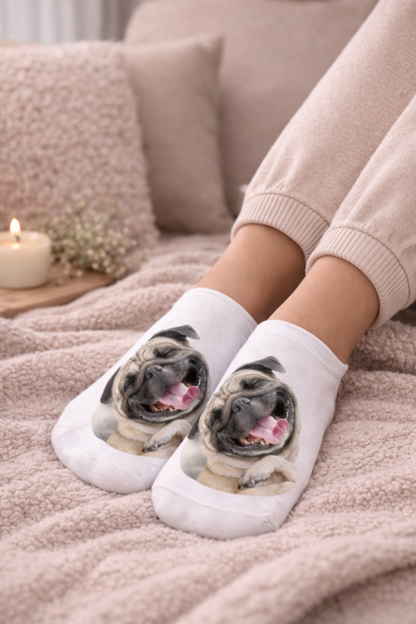 White ankle socks with printed pug dog faces, worn by a person sitting on a soft beige blanket with pillows and a candle in the background.