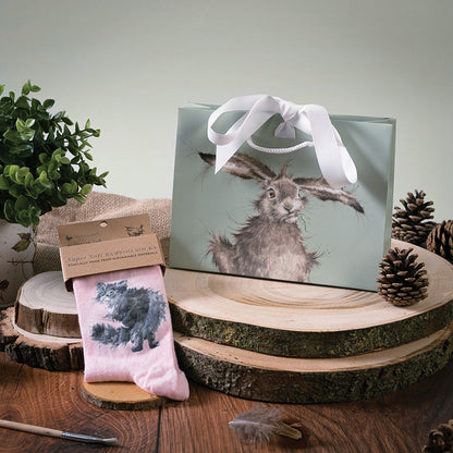 A gift set featuring a sage green haring bag with a white ribbon and a pink sock with a black cat illustration, displayed on rustic wood slices with pinecones and a small potted plant.