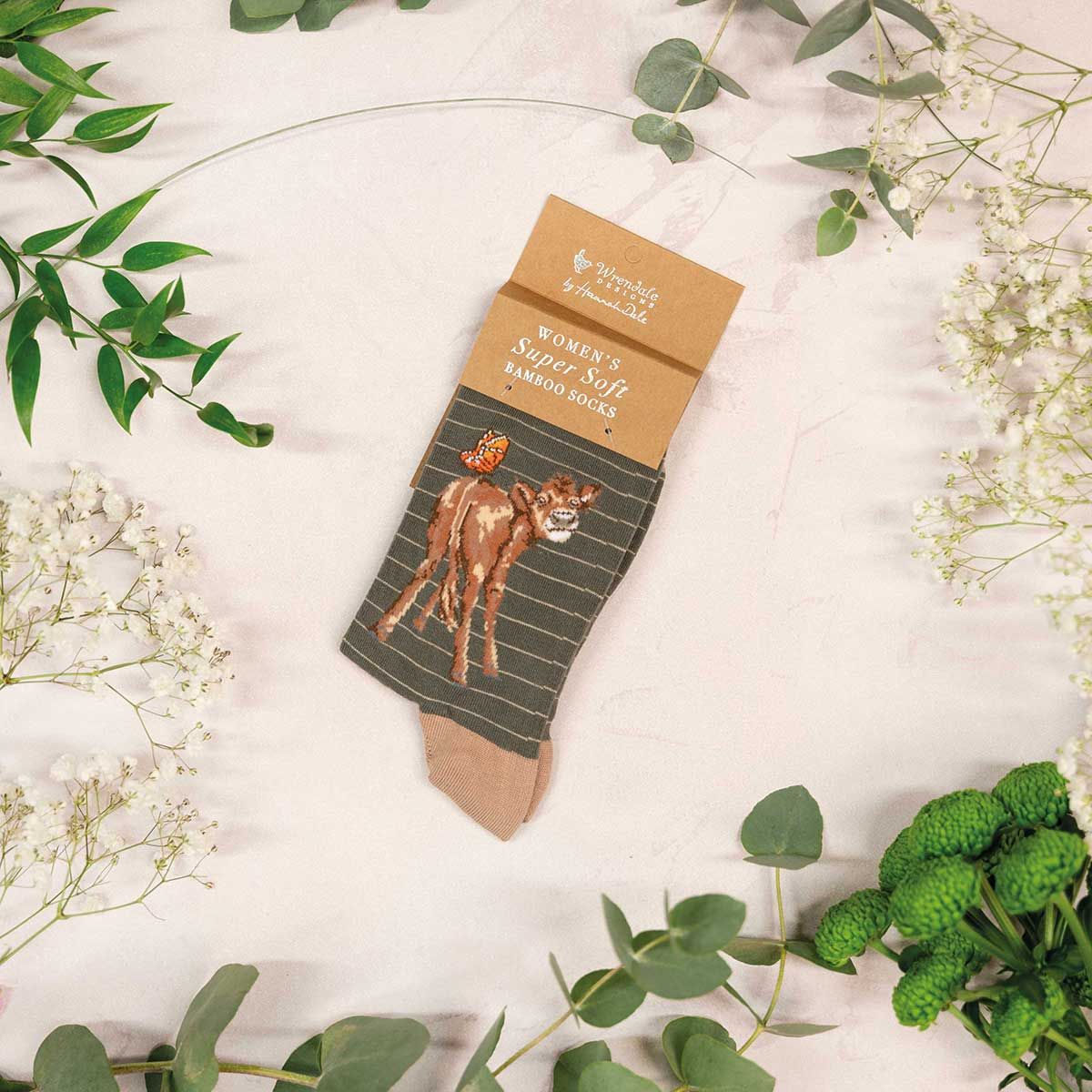 Sock with a cow design on a cardboard backing, surrounded by greenery and flowers.