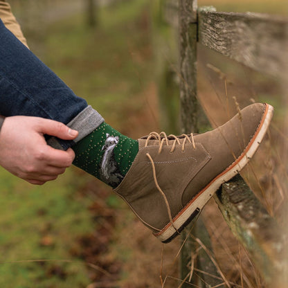 A person's foot wearing a brown suede lace-up boot and a green patterned sock with a gray animal design, resting on a weathered wooden fence outdoors.