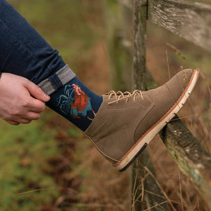 Person wearing a brown boot and colorful sock with a rooster design, standing on a wooden fence.