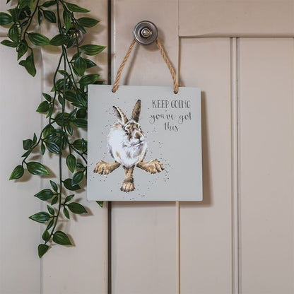 A square white wall hanging sign with a watercolor hare illustration and the words 'KEEP GOING you've got this,' suspended by a rope from a door knob against a cream paneled door with green trailing leaves on the left.