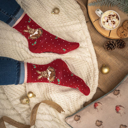 A pair of red Christmas socks with rabbit illustrations and small green holly accents, worn on a person's feet over a cream cable-knit blanket. A mug of hot chocolate, cookies, pinecones, and gold ornaments are arranged around the legs in a cozy flat-lay setting.