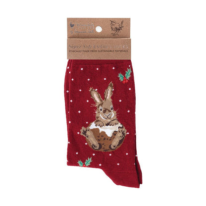 A pair of red Christmas socks with rabbit illustrations and small green holly accents, worn on a person's feet over a cream cable-knit blanket. A mug of hot chocolate, cookies, pinecones, and gold ornaments are arranged around the legs in a cozy flat-lay setting.