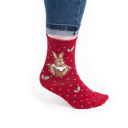 A single red crew sock worn on a foot, featuring a brown bunny holding a white gift box with green holly leaves and small white polka dots. The sock is shown on a person's foot with cuffed blue jeans against a plain white background.