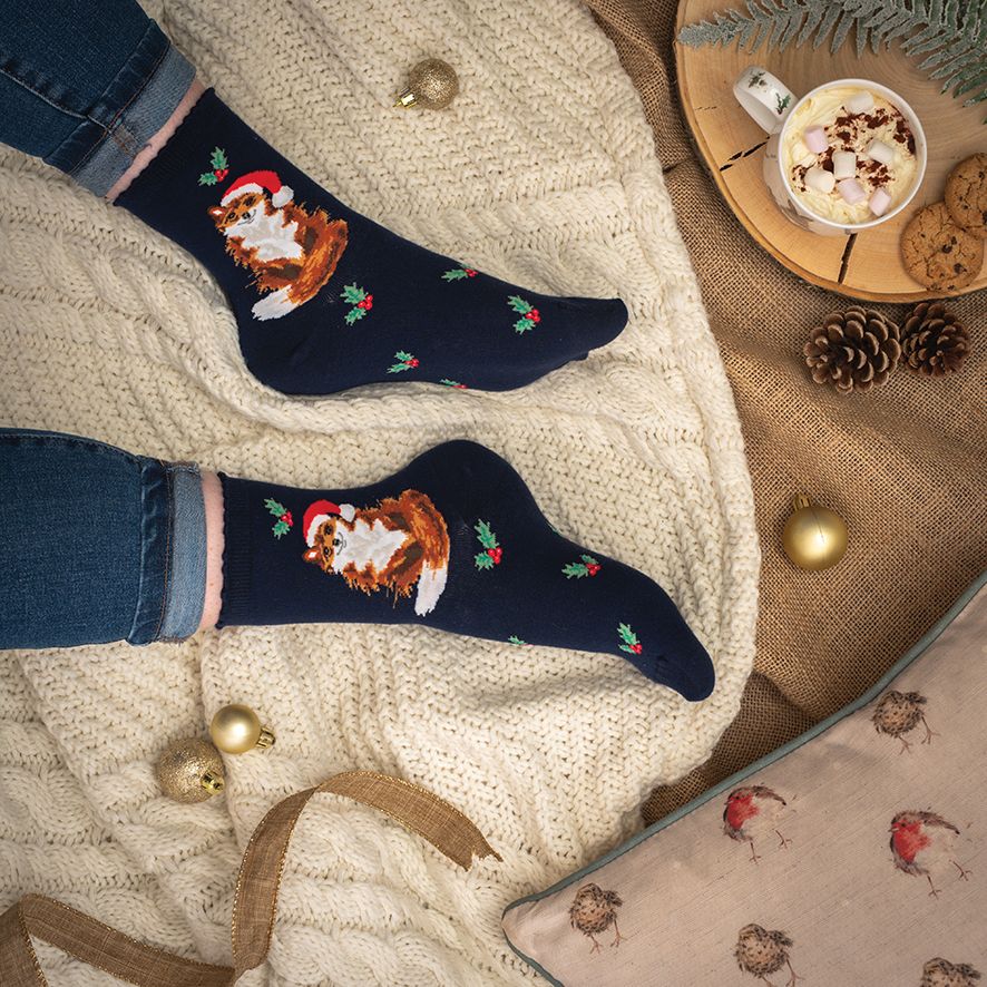 Navy blue socks with festive designs worn by a person, placed on a textured surface with a cup of hot chocolate and cookies.