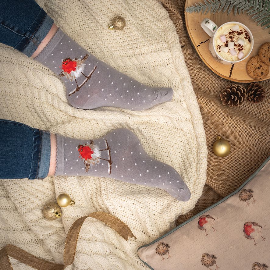 A pair of gray ankle socks with a white polka dot pattern and a red robin bird design, worn on feet resting on a cream cable-knit blanket. The scene is styled with blue jeans, a mug of hot chocolate, pinecones, gold ornaments, and a matching pillow.