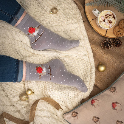 A pair of gray ankle socks with a white polka dot pattern and a red robin bird design, worn on feet resting on a cream cable-knit blanket. The scene is styled with blue jeans, a mug of hot chocolate, pinecones, gold ornaments, and a matching pillow.