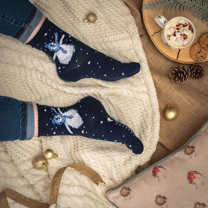 A pair of navy blue crew socks with a blue and white snowman design, worn on feet resting on a cream cable-knit blanket. The scene is styled with a cup of hot chocolate, pinecones, gold ornaments, and a cozy pillow.