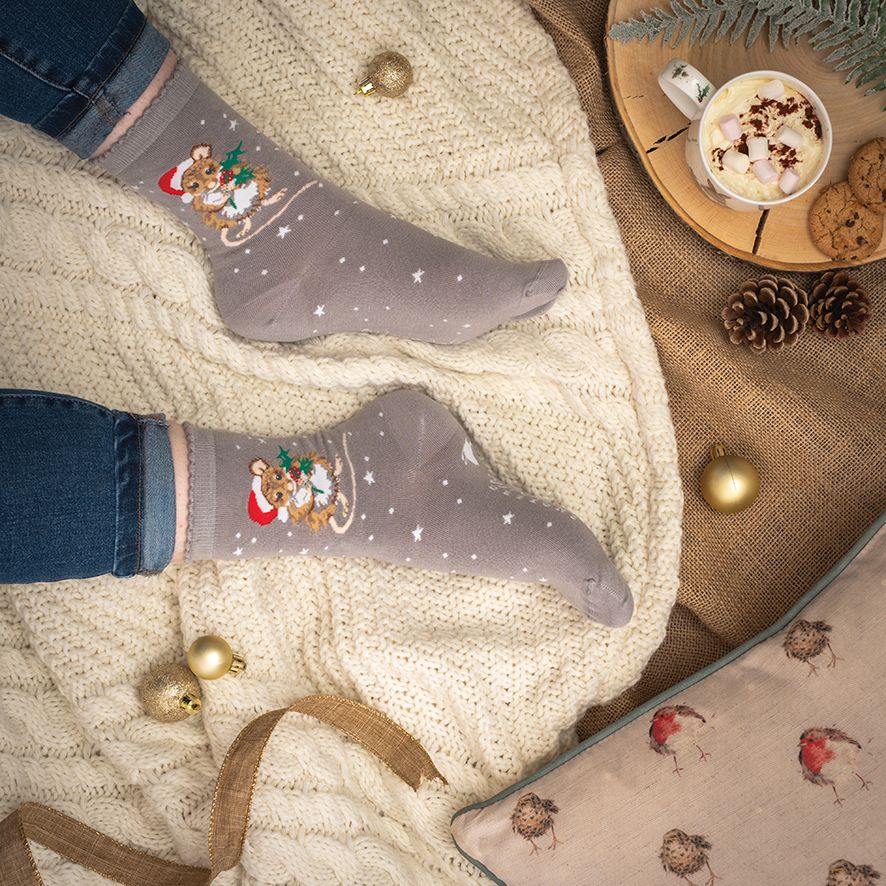 Person wearing festive socks with a Christmas design, surrounded by holiday decor including a mug of hot chocolate and cookies.