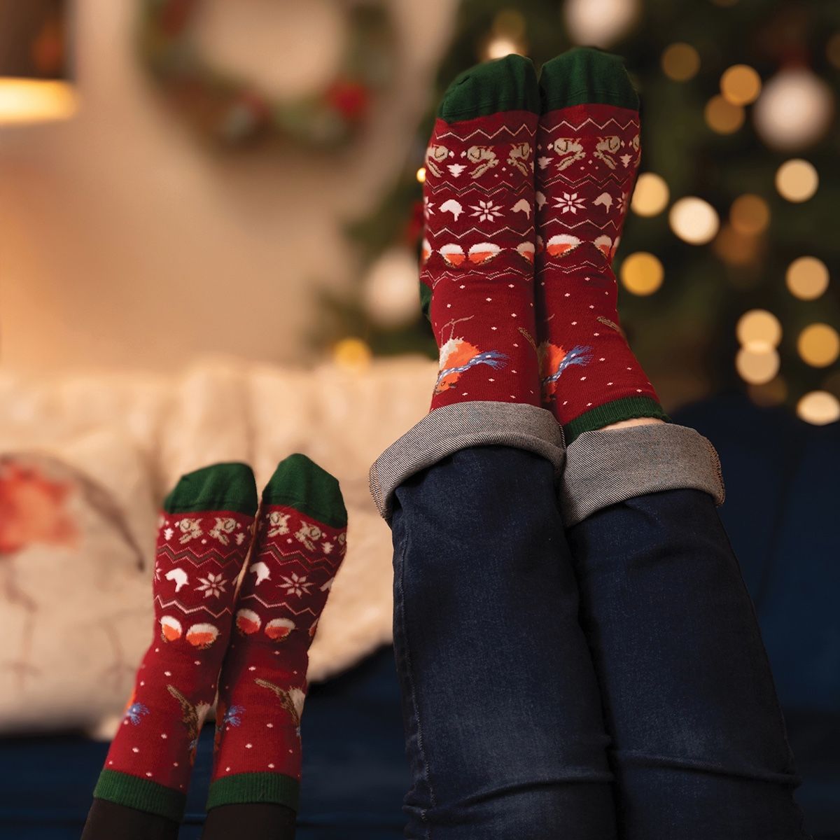 A pair of red Wrendale Designs Season's Tweetings Women's Christmas-themed crew socks with green cuffs and toes, featuring a festive fair isle pattern, worn on feet resting on a couch in a cozy living room setting with a blurred Christmas tree in the background.