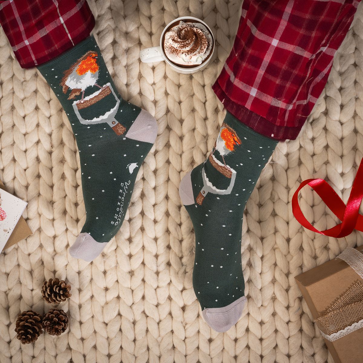 A pair of dark green Christmas-themed socks with a Santa Claus and hot cocoa design, shown on feet wearing red plaid pajama pants on a chunky knit blanket with a coffee cup and gift props.