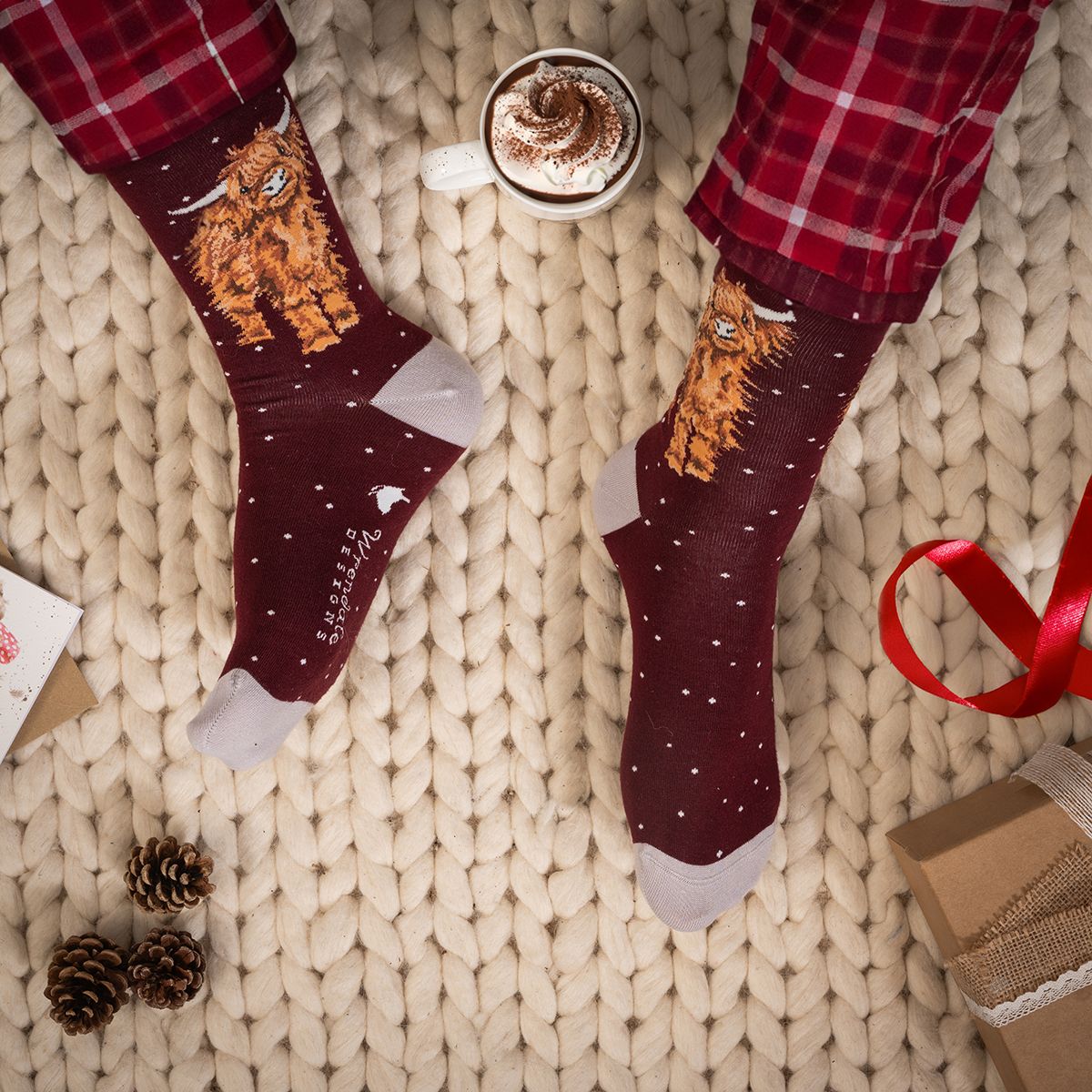 A pair of dark burgundy crew socks with a fluffy orange cat illustration and white speckles, worn with red plaid pajama pants on a beige knit blanket. A cup of hot cocoa, pinecones, and gift boxes are styled around the feet.
