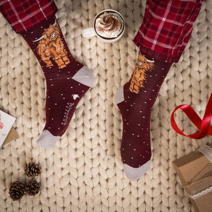 A pair of dark burgundy crew socks with a fluffy orange cat illustration and white speckles, worn with red plaid pajama pants on a beige knit blanket. A cup of hot cocoa, pinecones, and gift boxes are styled around the feet.