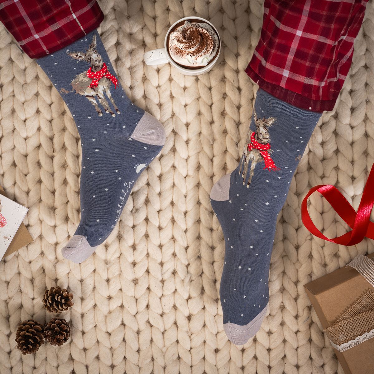 A pair of blue crew socks with a Christmas deer design, worn with red plaid pajama pants on a cozy beige knit blanket. A cup of hot chocolate, pinecones, and gift boxes are styled around the feet.