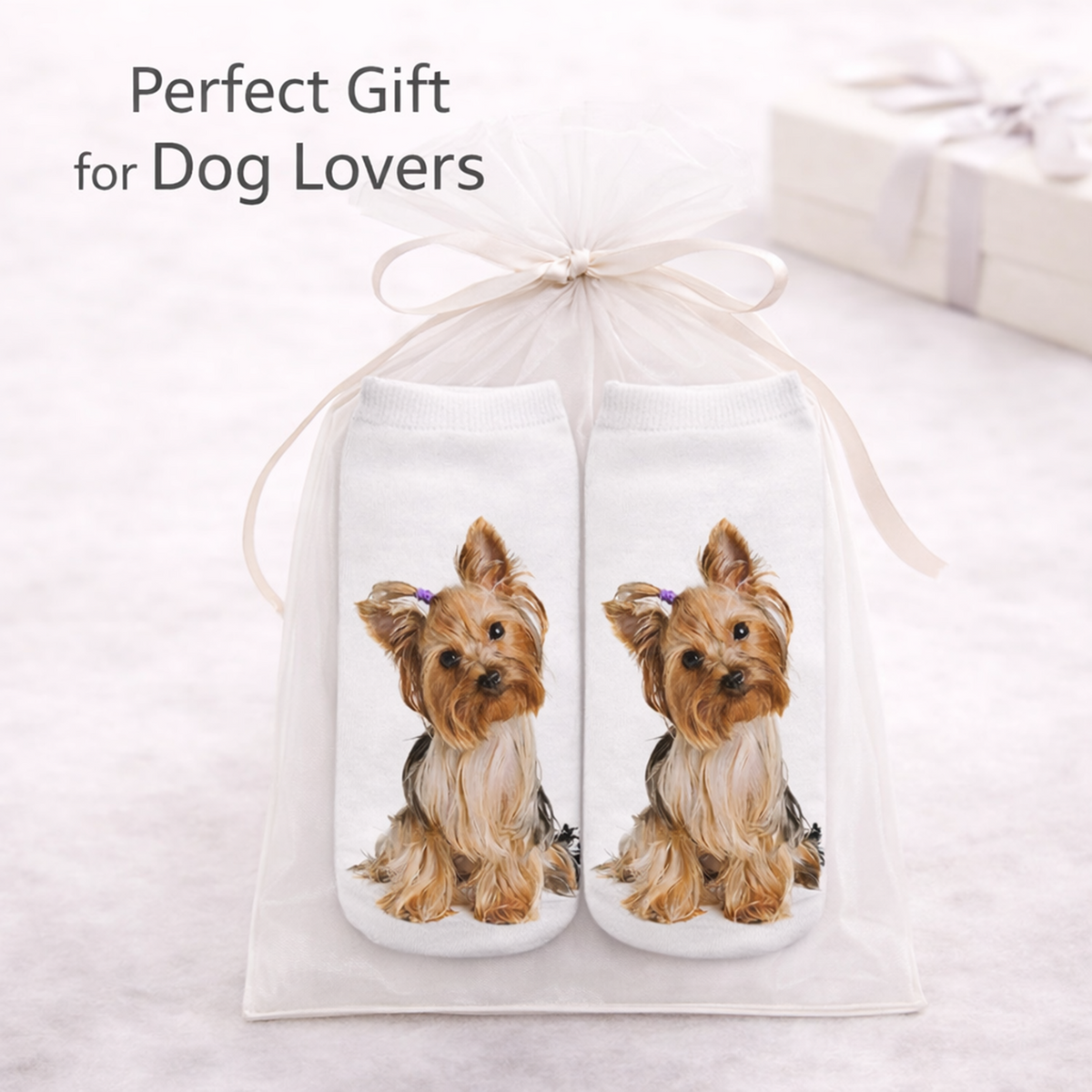 A gift set containing a pair of white trainer socks with printed illustrations of a Yorkshire Terrier, presented in a sheer white gift bag tied with a ribbon. The image includes the text 'Perfect Gift for Dog Lovers' and a blurred gift box in the background.