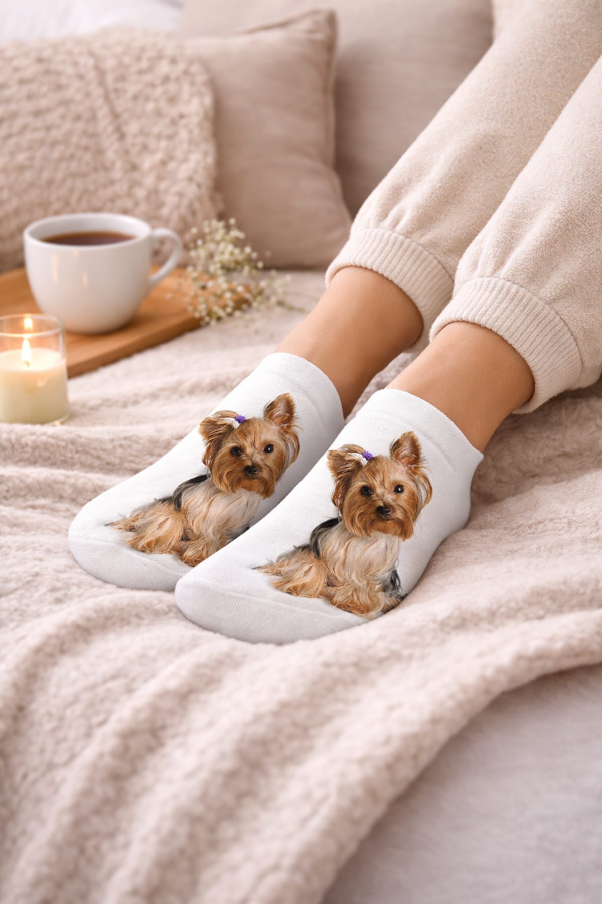 A person wearing white ankle socks with printed Yorkshire Terrier dog illustrations, sitting on a cosy beige blanket with a cup of coffee, candle, and pillows in the background.